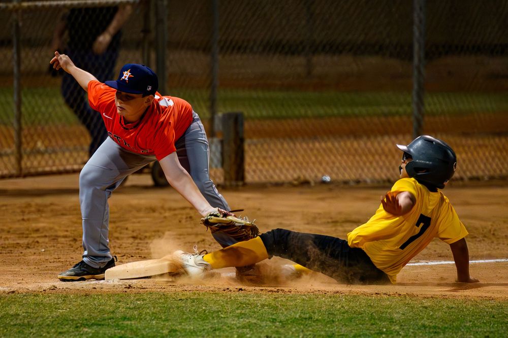 Youth baseball player in orange jersey tags out sliding runner in yellow uniform at base.