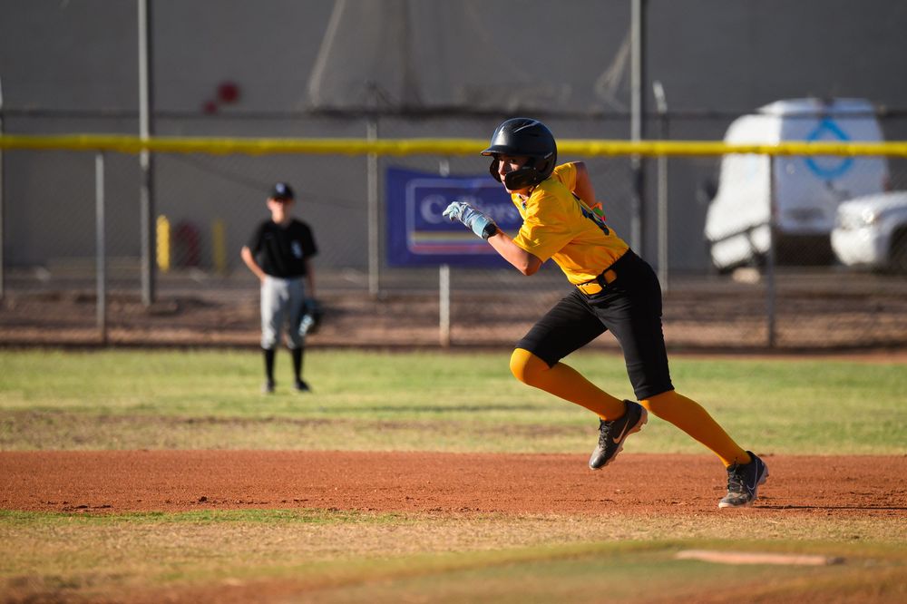 Youth softball player in yellow uniform sprints between bases during an outdoor daytime game.