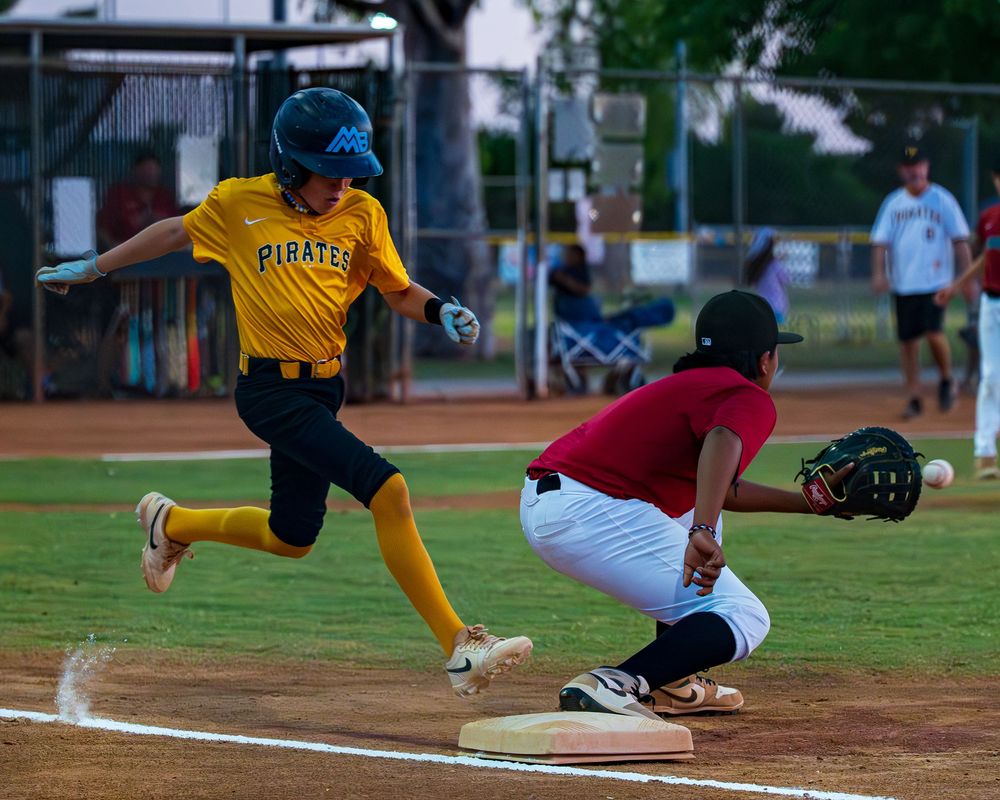 Young Pirates baseball player runs to first base as fielder attempts to catch the ball during a youth league game.
