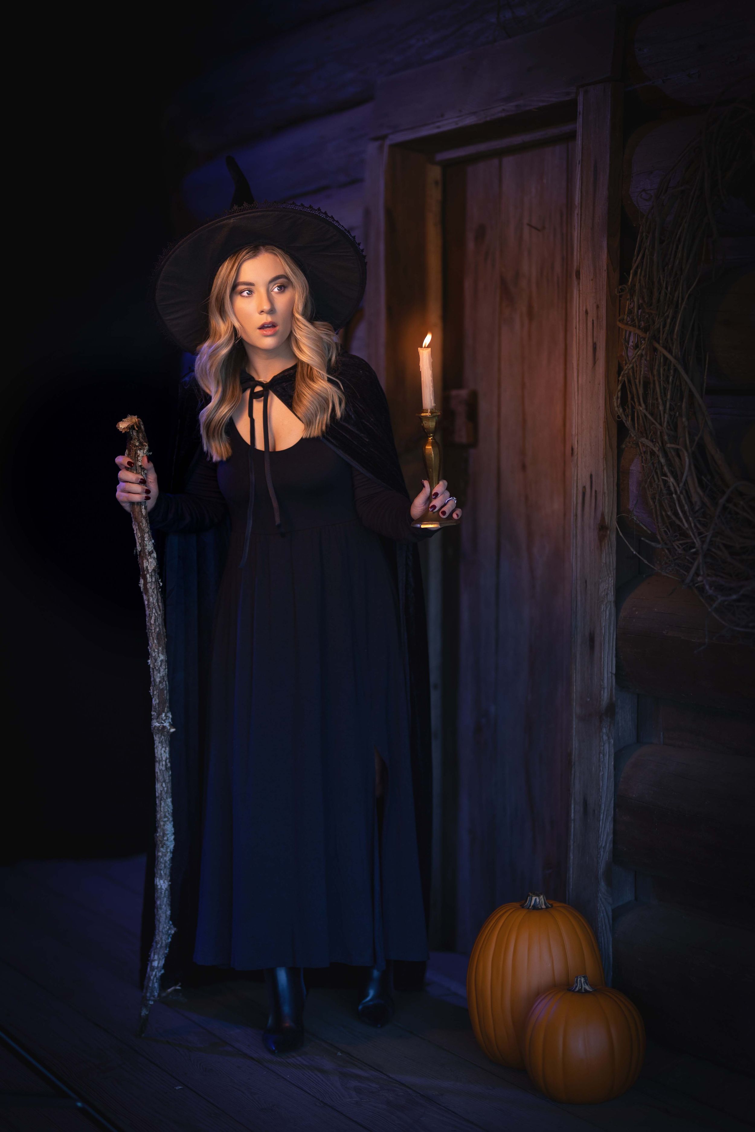A witch in a black dress and hat stands at a doorway holding a staff and candle with pumpkins on the ground at night.