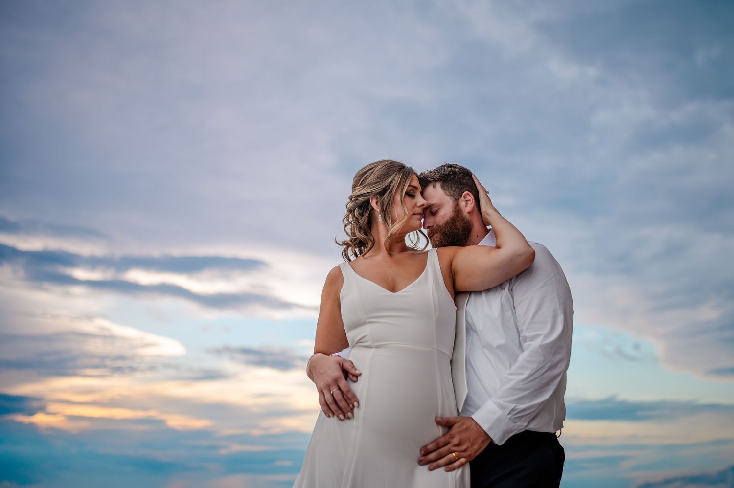 A romantic couple embraces against a dramatic sunset sky with soft clouds in the background.