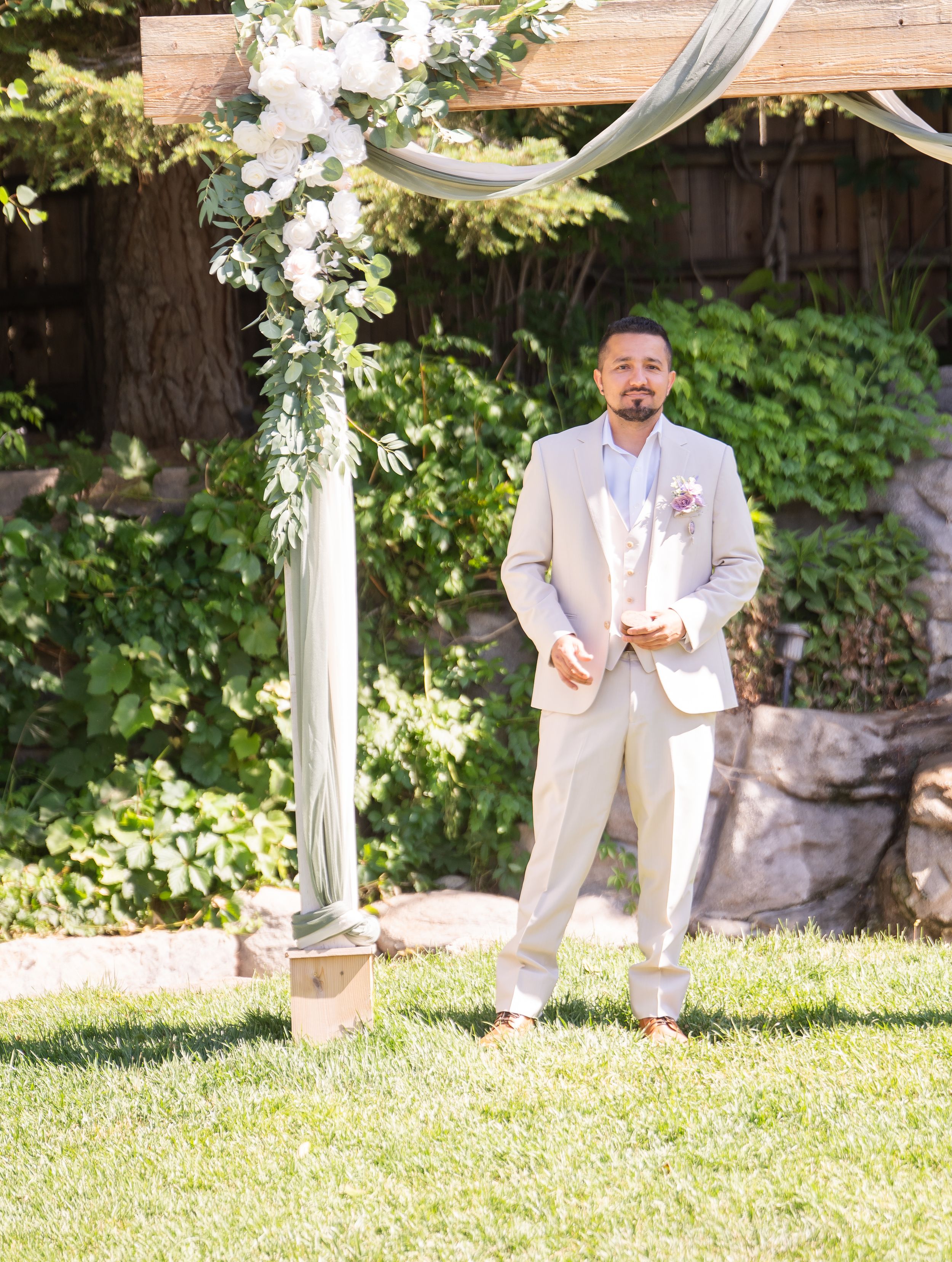 A person in a light beige suit stands under a white floral arch decorated with fabric draping in a garden setting.