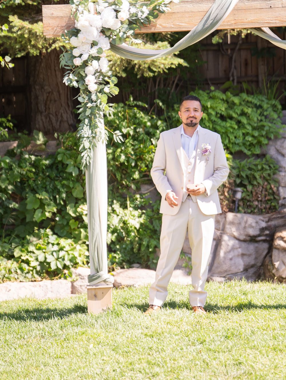 A person in a light beige suit stands under a white floral arch decorated with fabric draping in a garden setting.