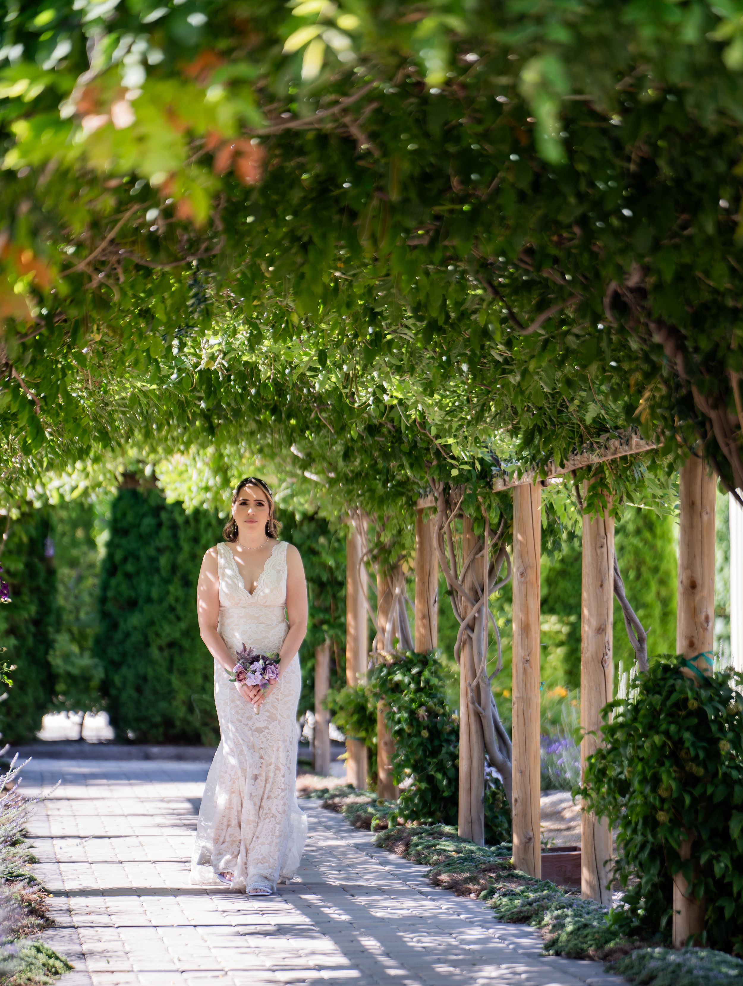 A bride in a lace gown walks along a garden path under a vine-covered pergola on a sunny summer day.