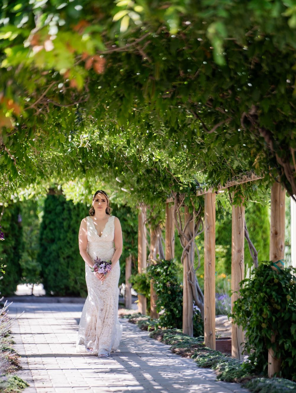 A bride in a lace gown walks along a garden path under a vine-covered pergola on a sunny summer day.