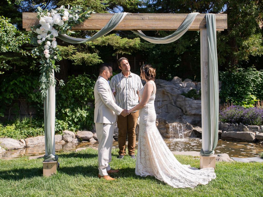 Elegant outdoor wedding ceremony setup with white draped archway and decorative accents against lush green landscape.