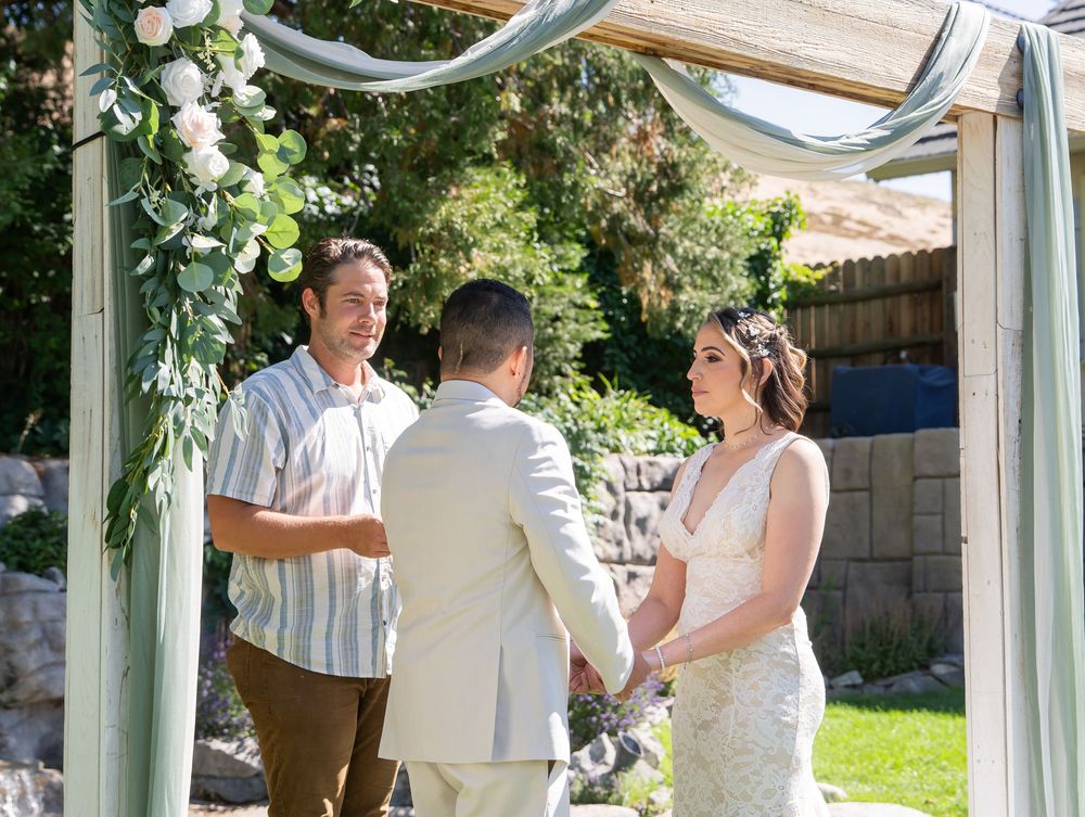 A couple stands beneath a white draped wedding arch adorned with greenery during an outdoor backyard ceremony.