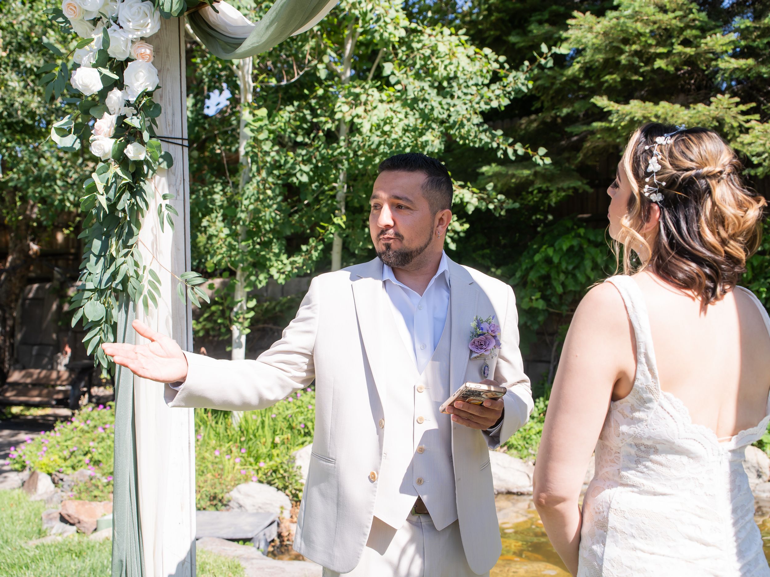 A bride and groom share a candid moment under a floral arch during an outdoor garden wedding ceremony.
