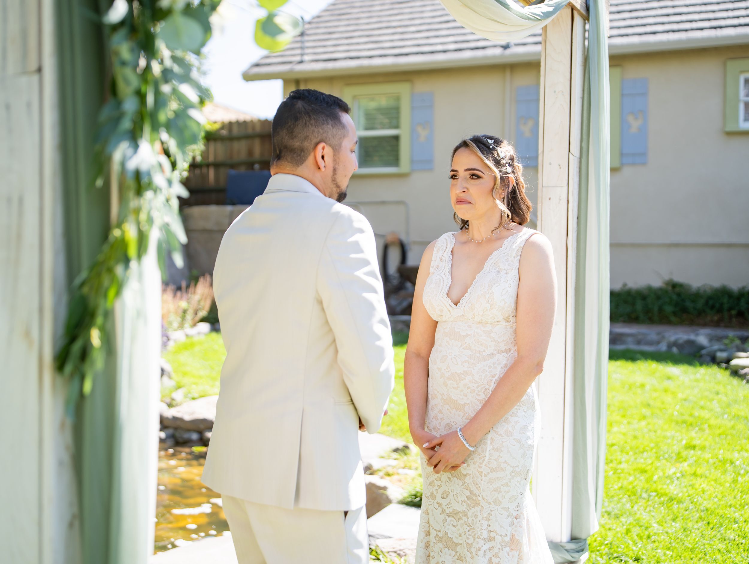 A bride and groom share a moment in white attire during their first look outside on a sunny garden lawn.
