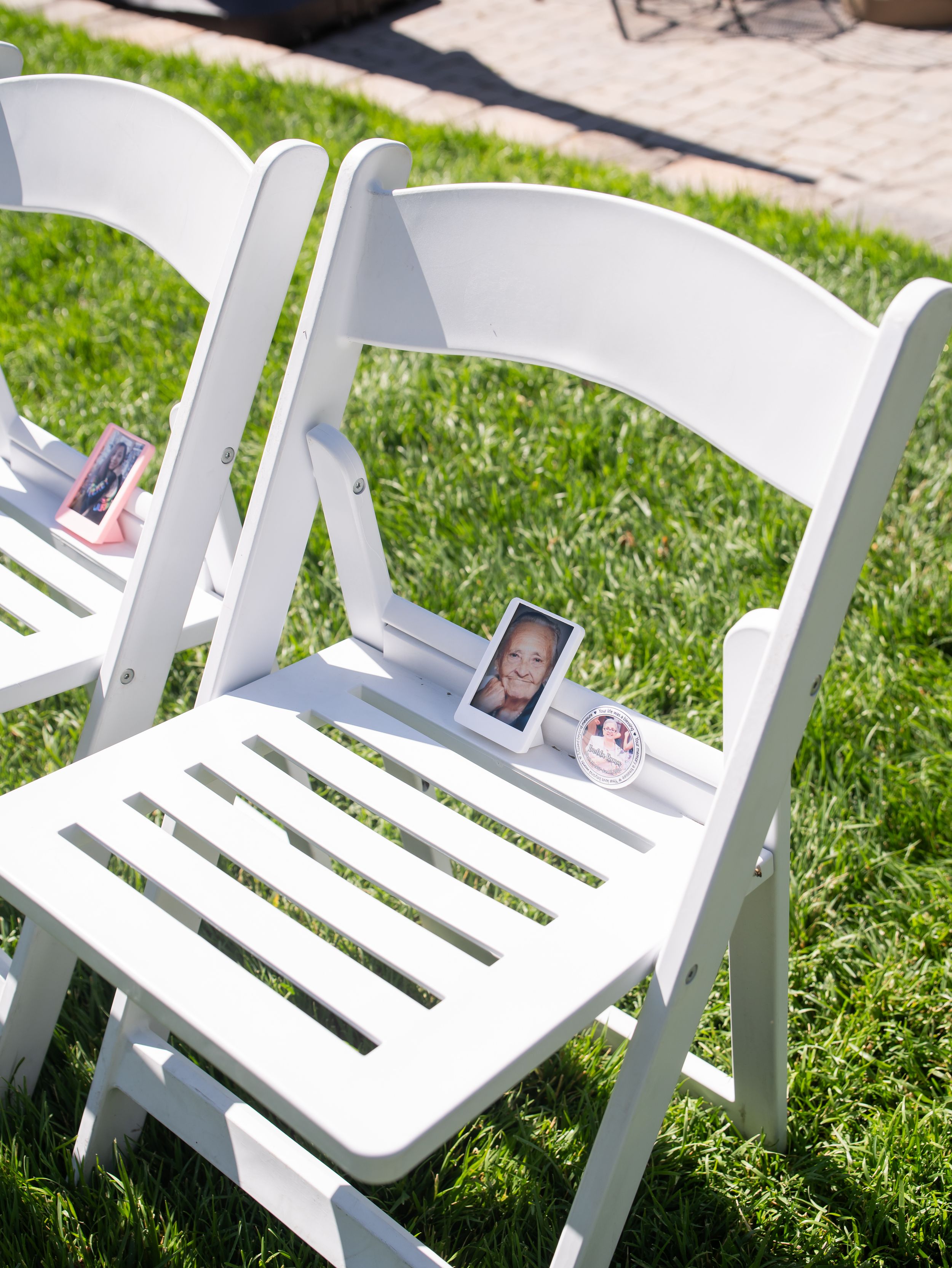 White folding chairs arranged on grass with cell phones placed on seats for wedding guests.