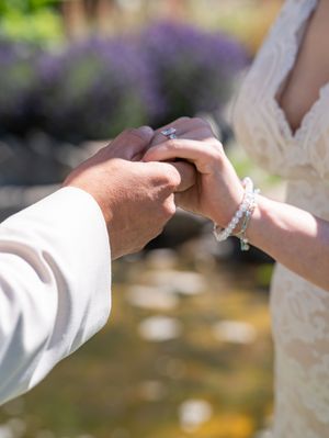 Close-up photograph of hands exchanging a wedding ring during an outdoor ceremony with flowers in background.