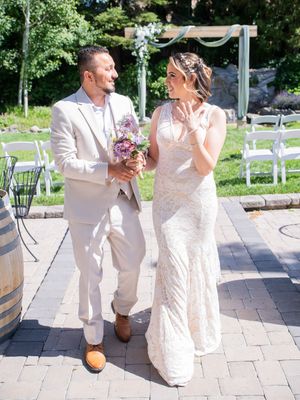 A couple walks joyfully together at an outdoor wedding ceremony featuring a decorated arch and white chairs on a brick patio.