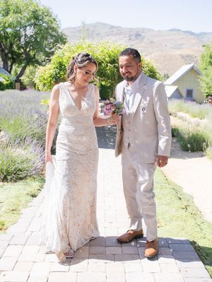 Couple in matching white attire walking together through a lavender garden path with mountains in background.