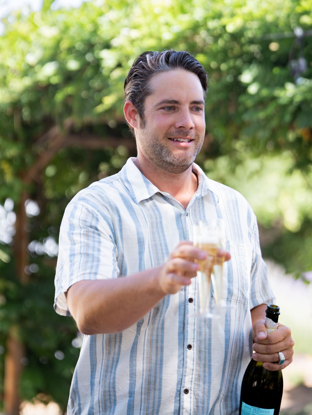 Person in striped button-down shirt holding champagne bottle and glass outdoors on sunny day.