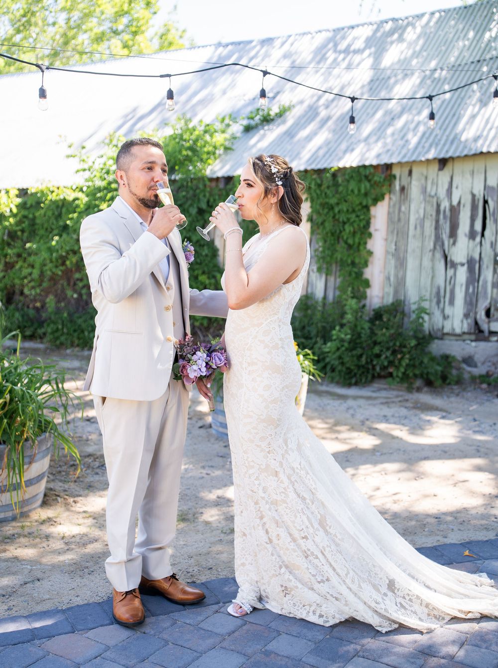 A romantic wedding photo outdoors with string lights and rustic barn backdrop featuring a couple in cream and lace attire.