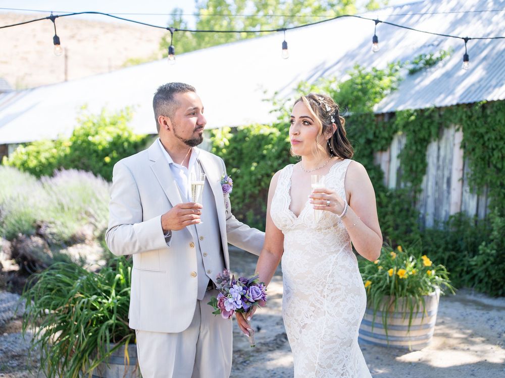 A couple shares a candid moment laughing and holding drinks during a rustic outdoor wedding celebration.