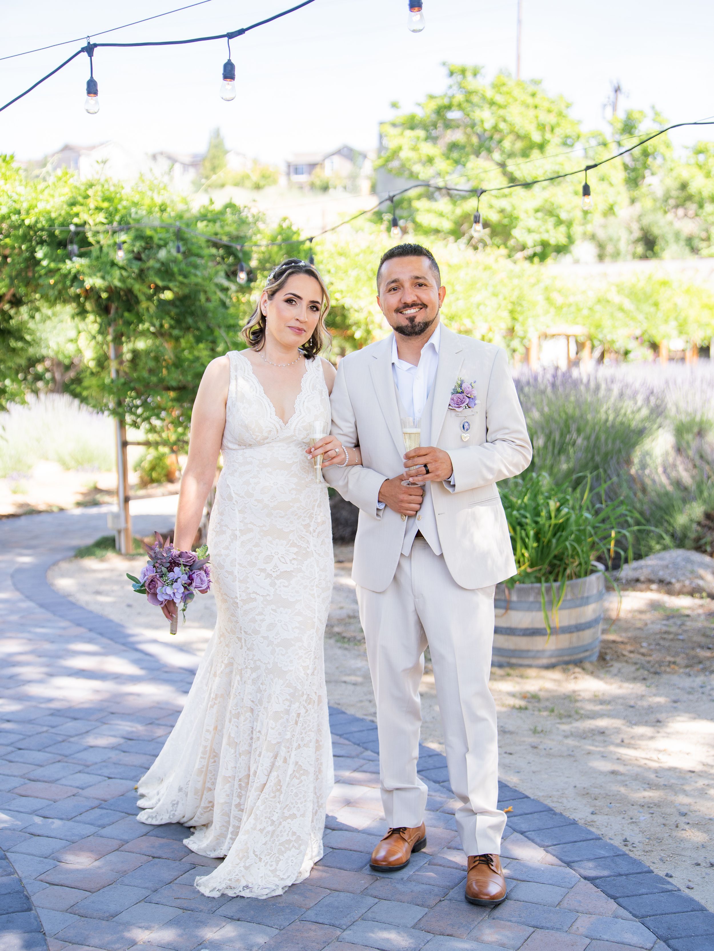 A couple poses on a stone path surrounded by lush garden landscaping, string lights overhead at an outdoor wedding venue.