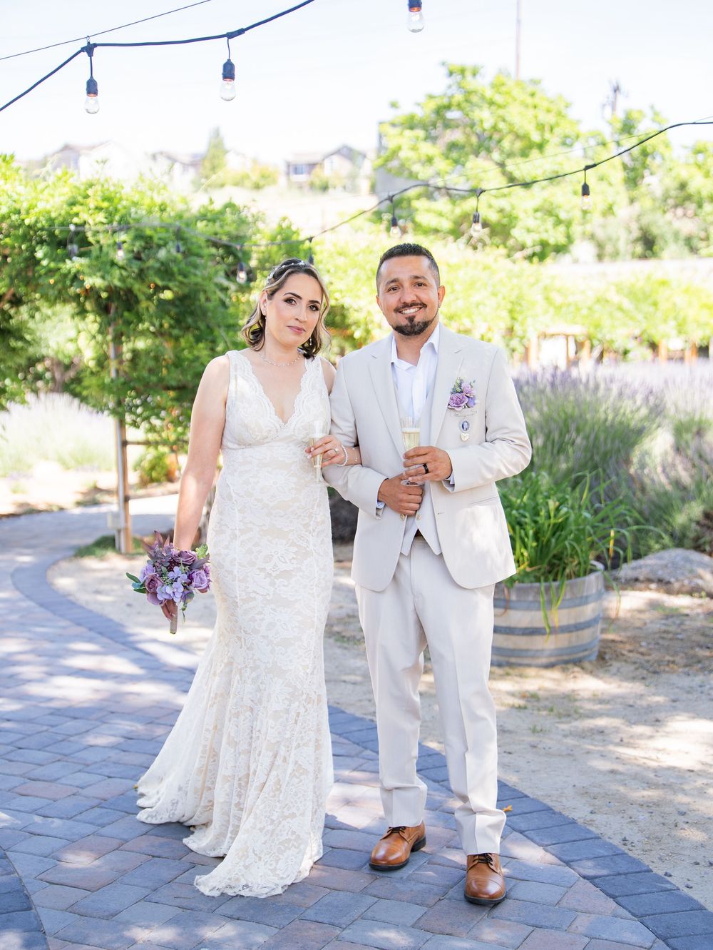 A couple poses on a stone path surrounded by lush garden landscaping, string lights overhead at an outdoor wedding venue.