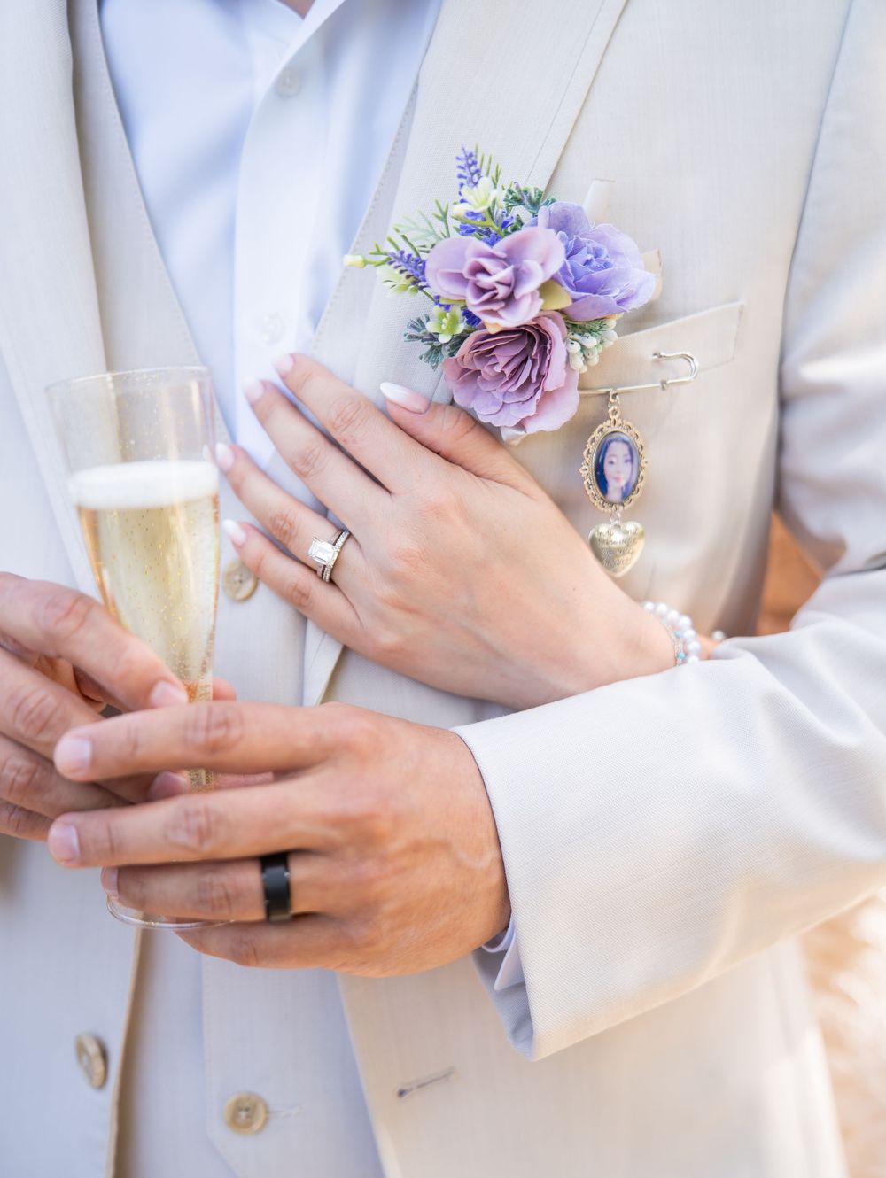 Wedding detail photo showing hands with rings, champagne glass, and purple floral boutonniere against cream suit.