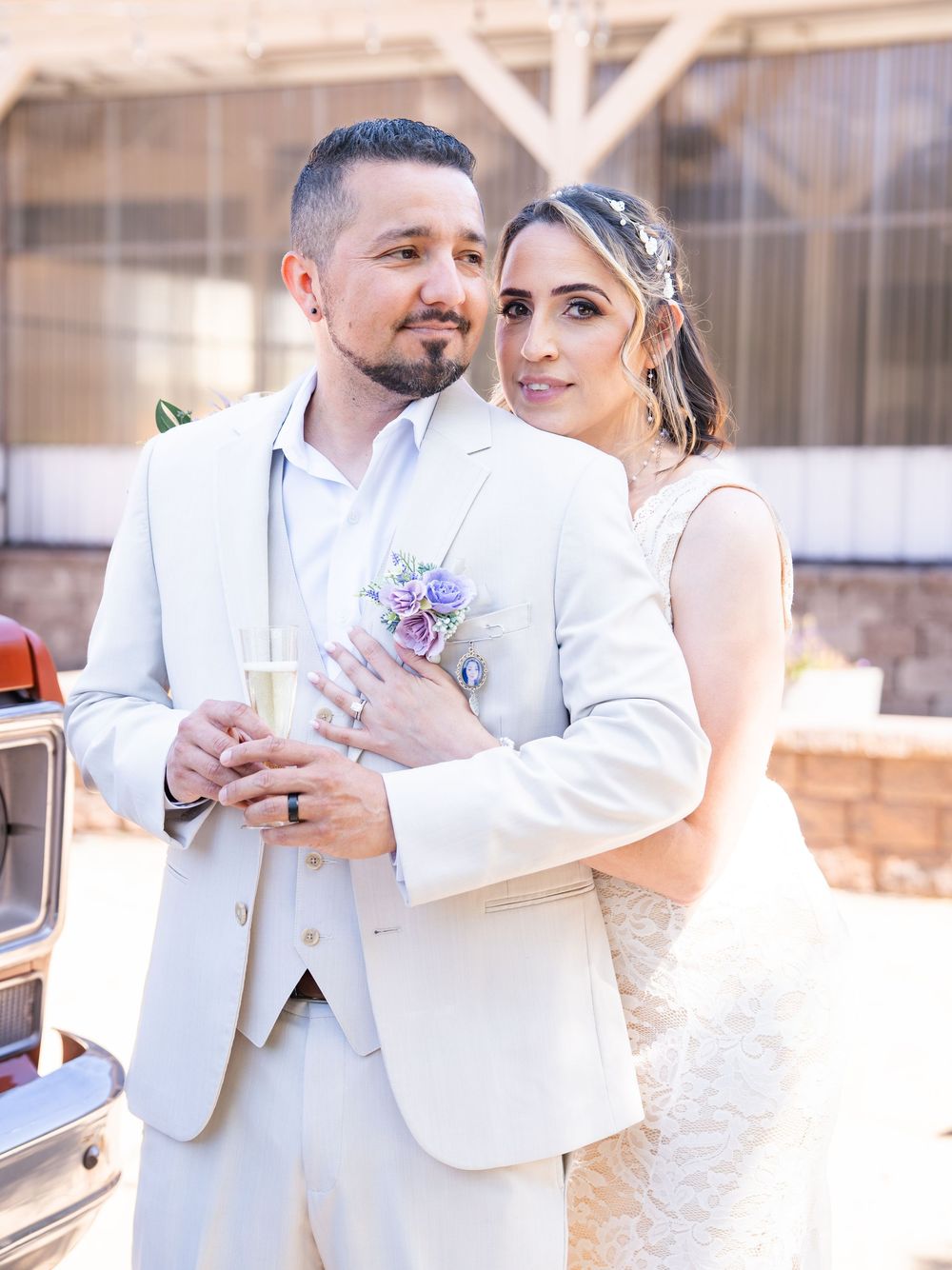 A couple in white wedding attire embraces while holding champagne glasses during their celebration.