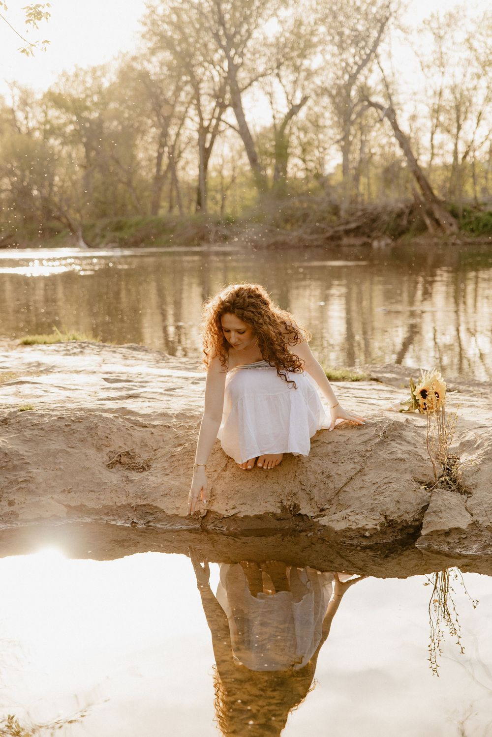 A person in white perches on a rock overlooking a tranquil river at golden hour in Frederick Maryland