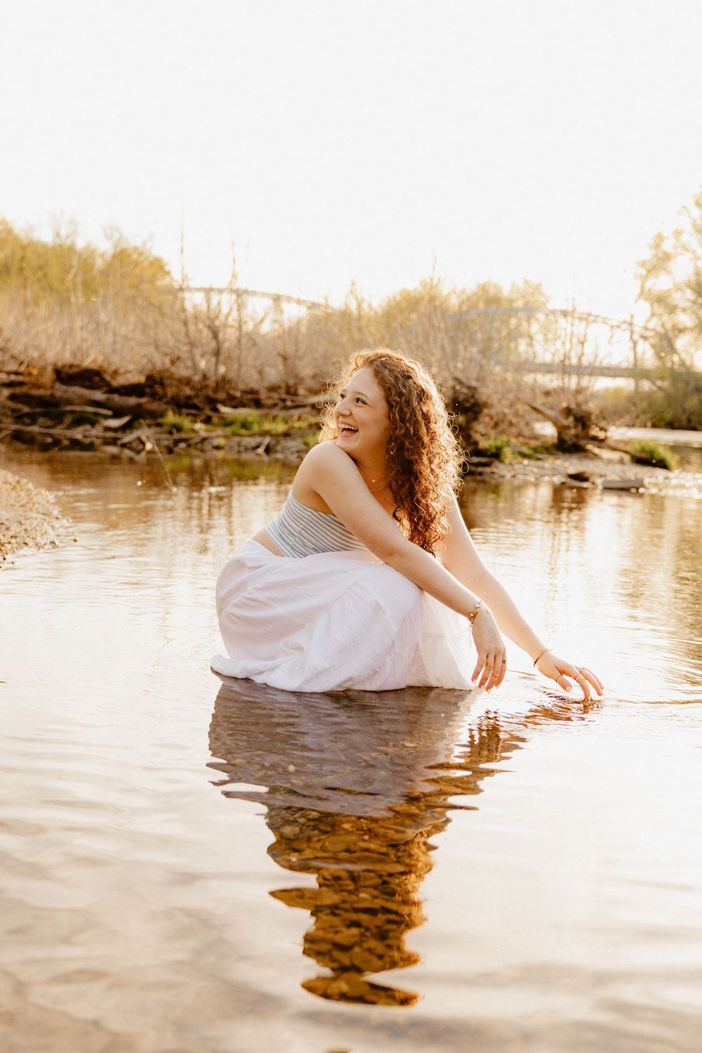 A person in white dress touches water while sitting on rocks by a serene river at sunset.