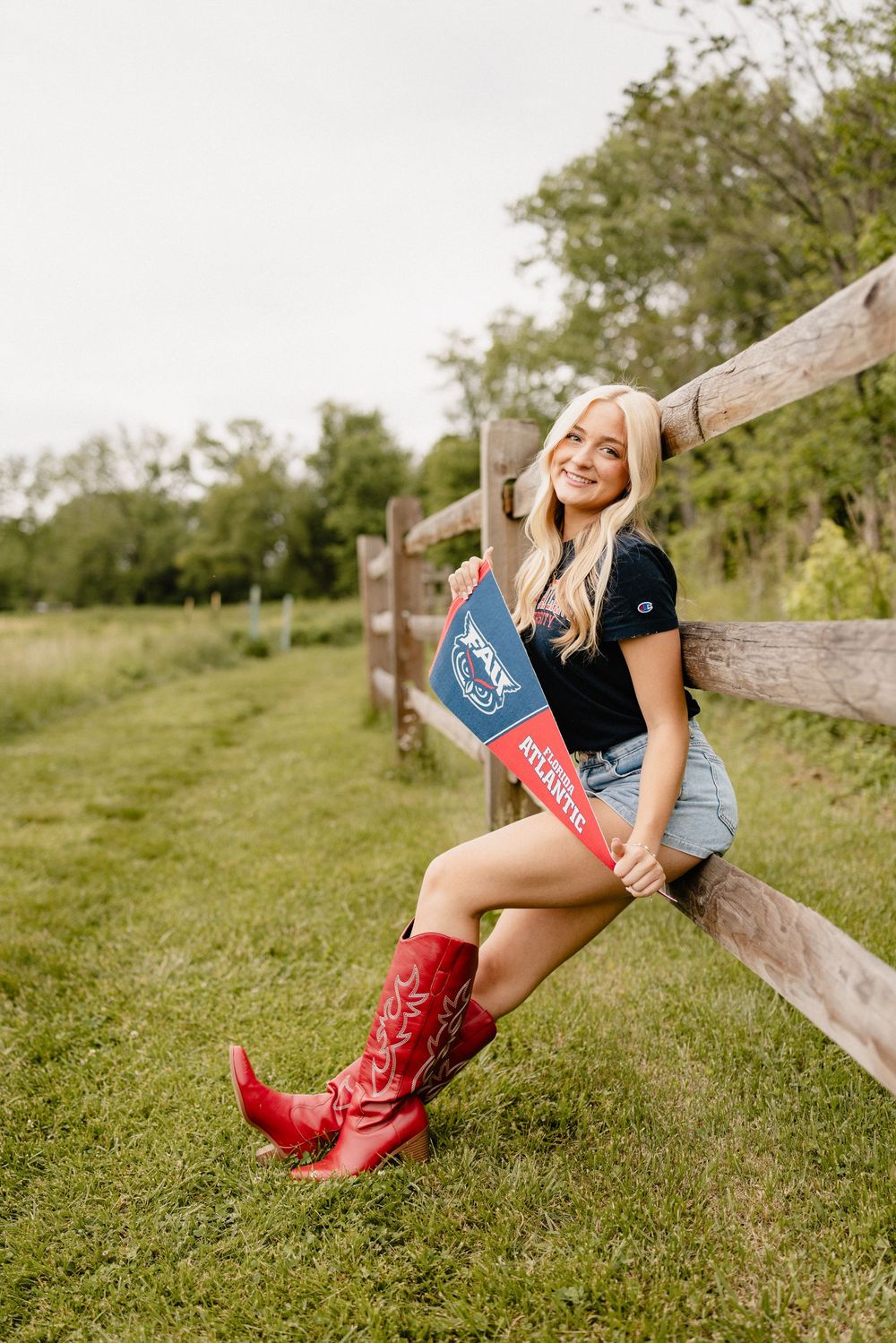 A person in red cowboy boots leans against a wooden fence in a pastoral countryside setting.