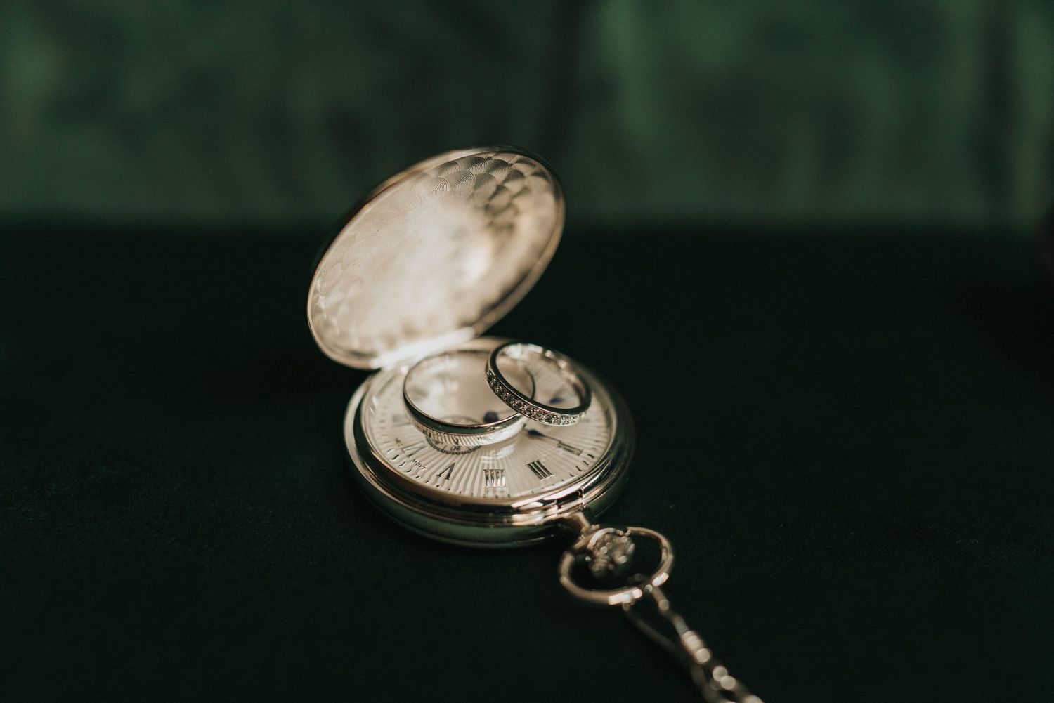 Vintage gold pocket watch with chain lies open against dark green background, with wedding rings sat on top.