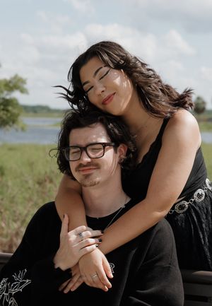 A romantic couple dressed in black embraces while sitting in a grassy field on a sunny day.