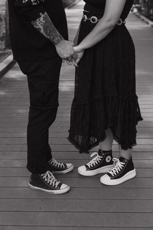 Black and white photo showing couple's feet in matching Converse sneakers on wooden deck.