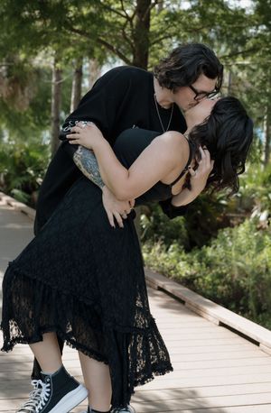 Couple in black clothing shares intimate moments on a wooden boardwalk surrounded by greenery.