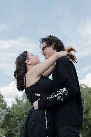 Couple in black attire shares romantic moments against cloudy sky.