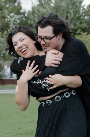 Couple in black clothing laughs and embraces in an outdoor setting.