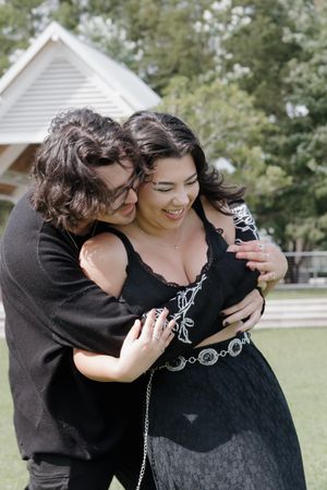 A romantic couple embraces outdoors in front of a white house on a sunny day.