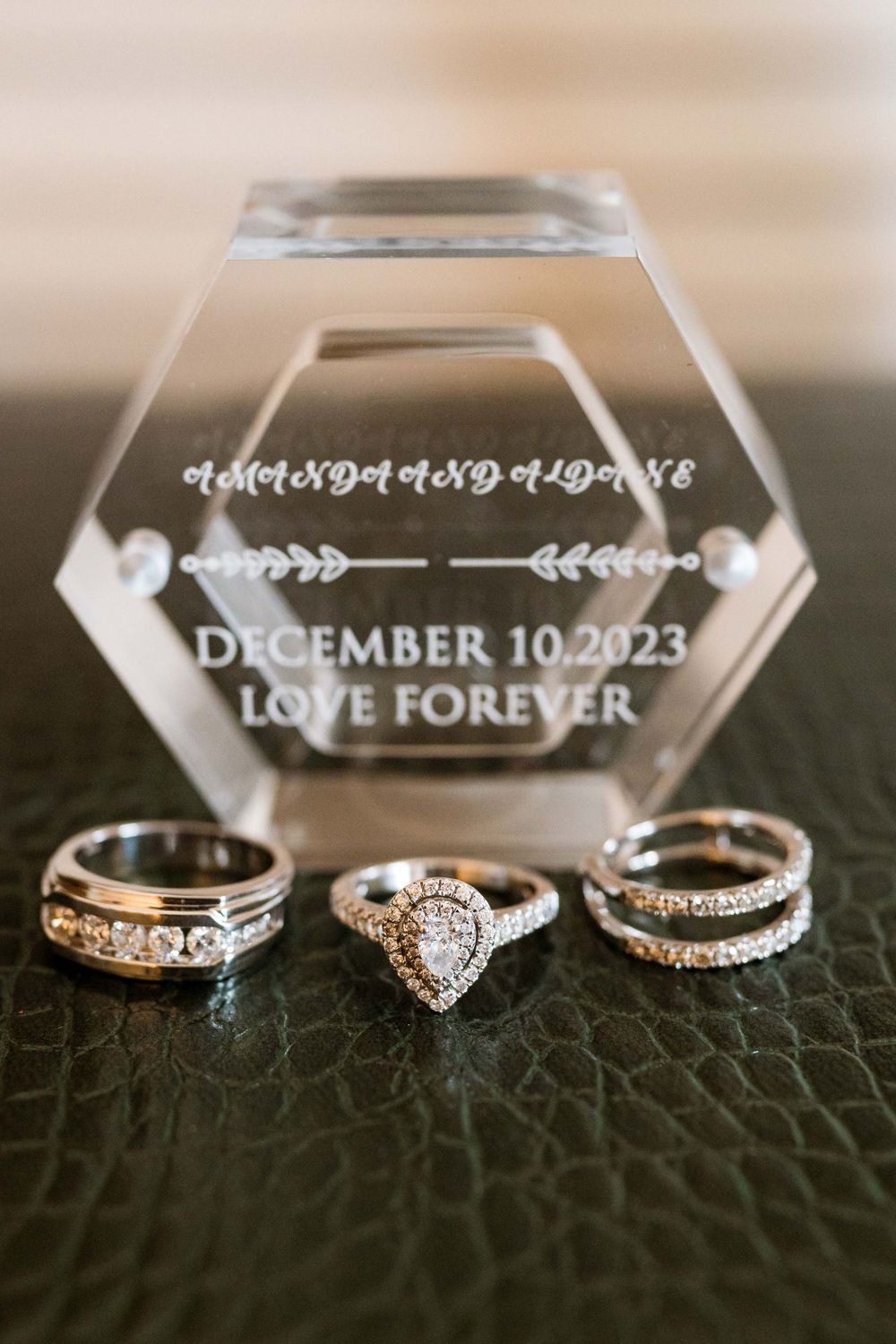 Wedding rings and crystal hexagonal display with December 10 2023 date engraving on textured dark surface.