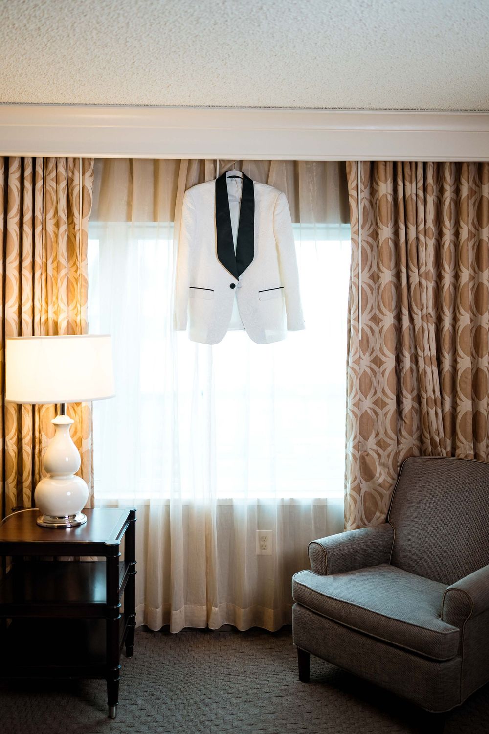 An elegant white tuxedo jacket hangs in a hotel room with beige curtains and a grey armchair.