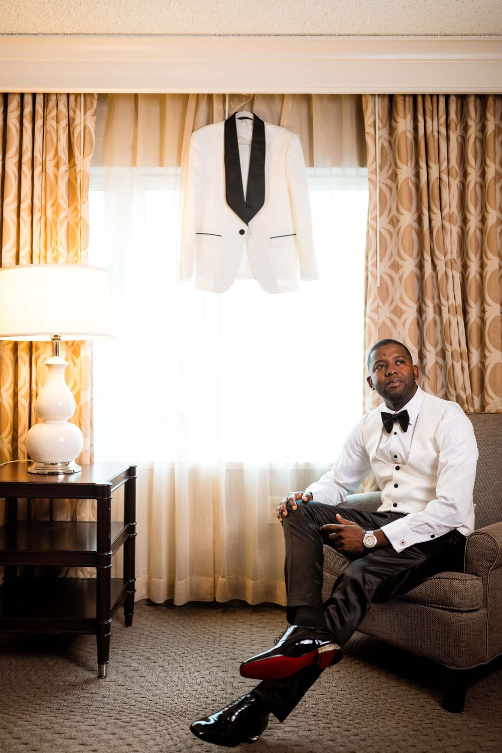 Elegant formal wear setup featuring a white tuxedo jacket hanging near a window and a person seated in a hotel room.