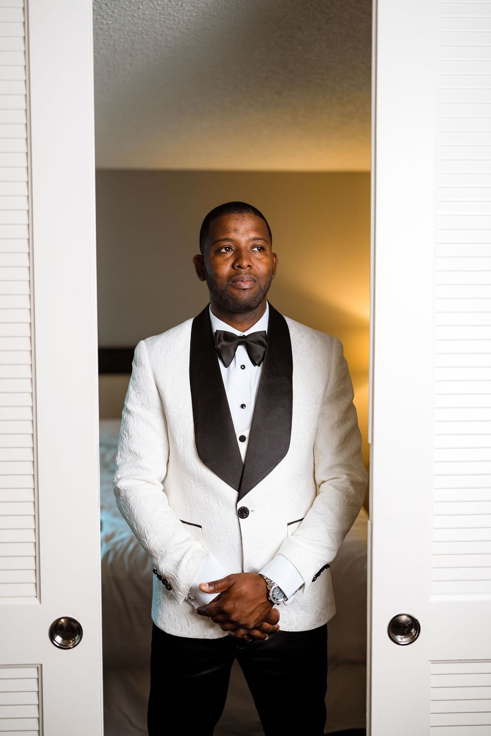 A formal white tuxedo jacket with black bow tie is shown in a doorway with warm lighting.