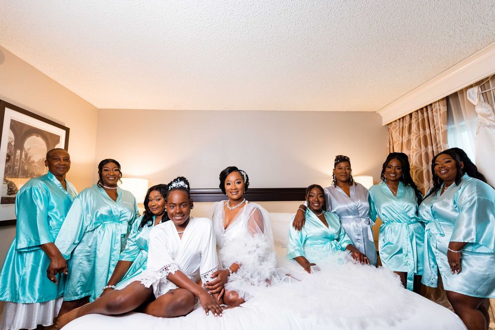 Group wearing matching turquoise silk robes and white outfits pose together on hotel bed during celebration.