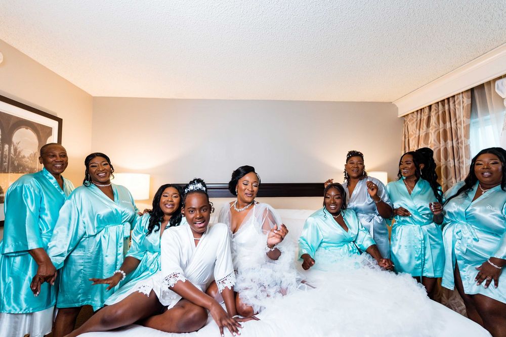 A bridal party wearing turquoise silk robes poses together on a bed in a hotel room during pre-wedding preparations.