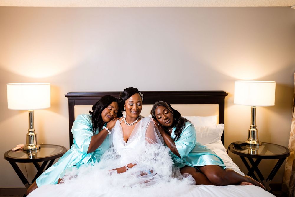 A bride in white dress poses on bed with two bridesmaids wearing turquoise robes and smiling warmly.