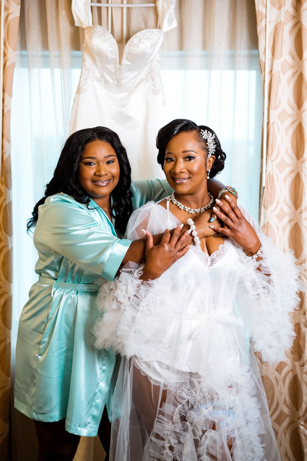 A bride in a flowing white gown poses with someone in a mint silk robe beside a hanging wedding dress.
