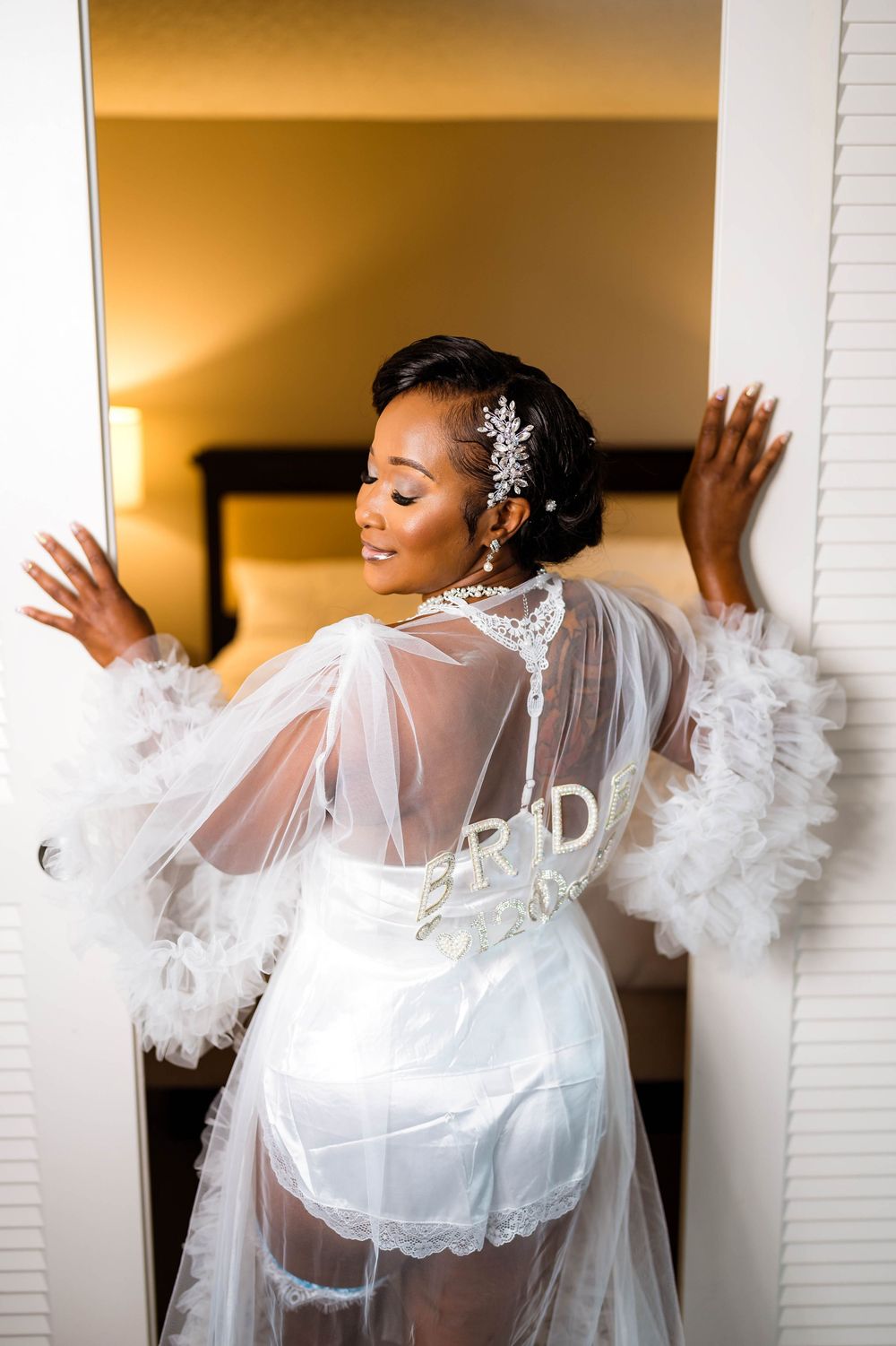 Bride in sheer white robe with feather details poses playfully against white doors on her wedding day.