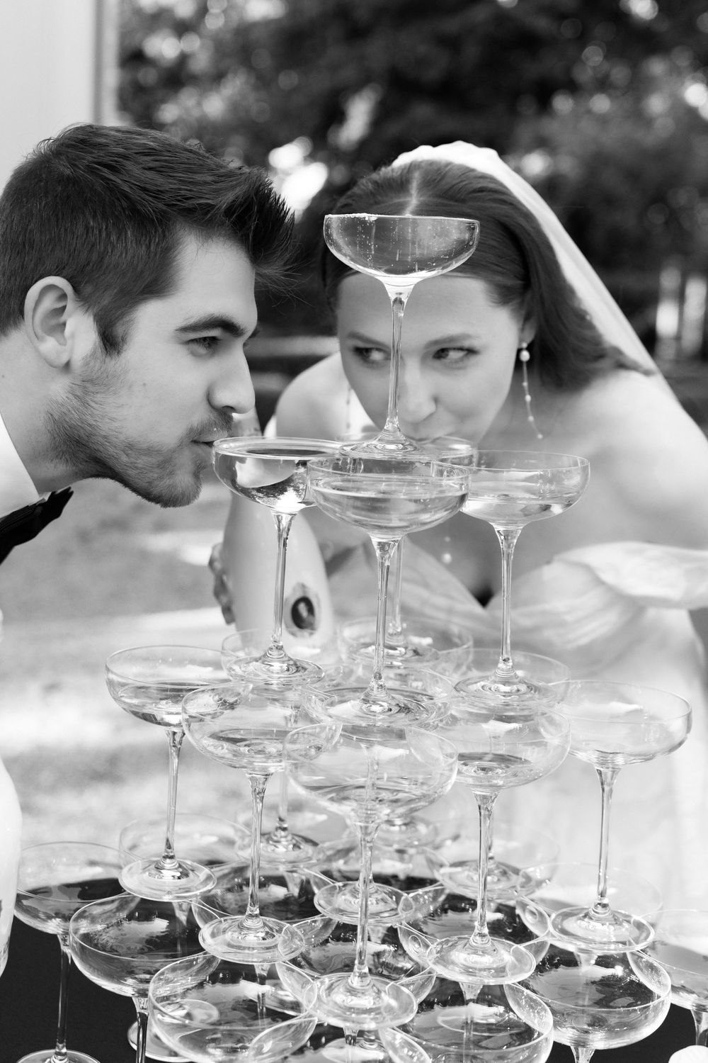 A bride and groom sip champagne from a tower of elegant coupe glasses at their wedding celebration.
