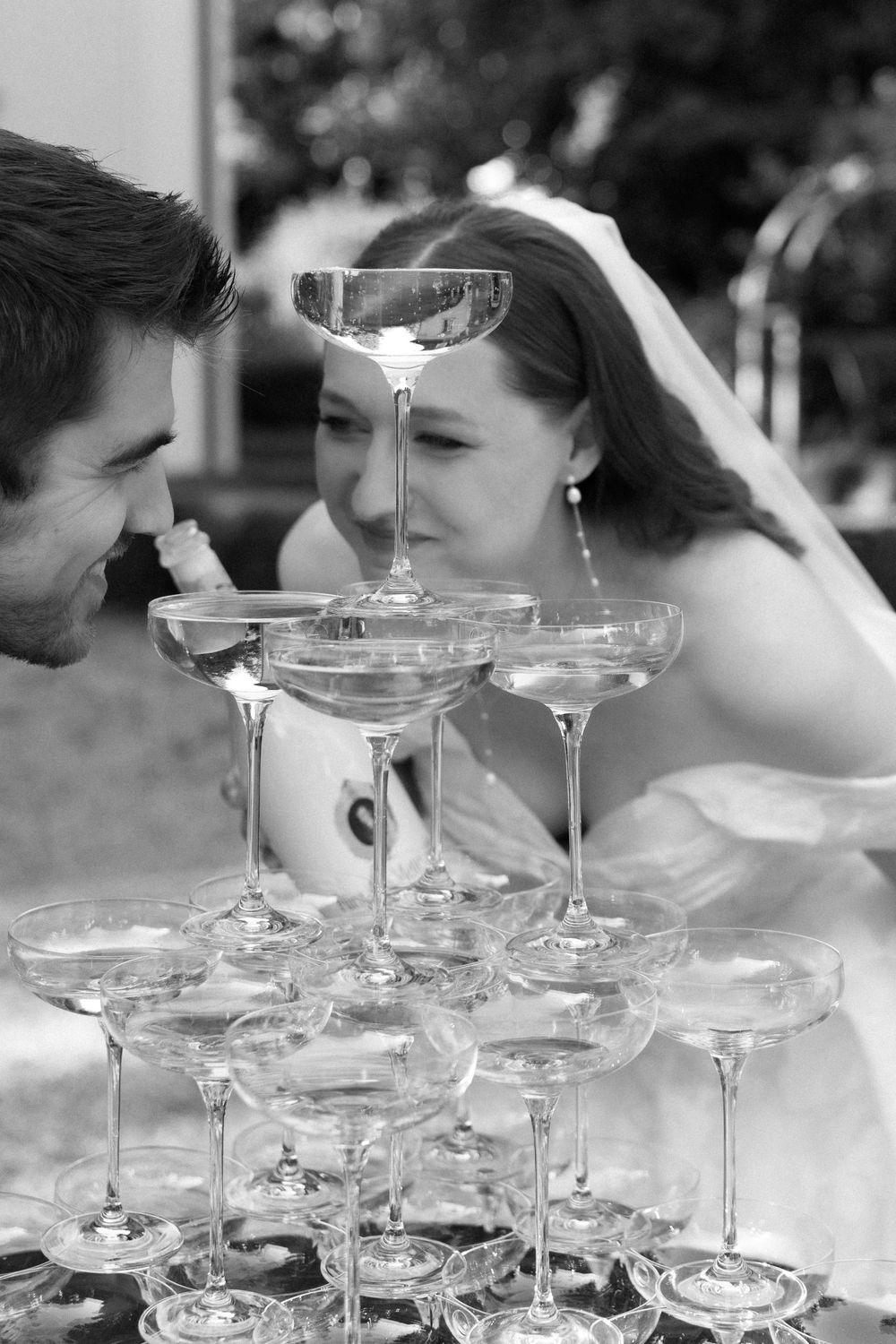 A black and white photo of a couple peering through a champagne tower at a wedding celebration.