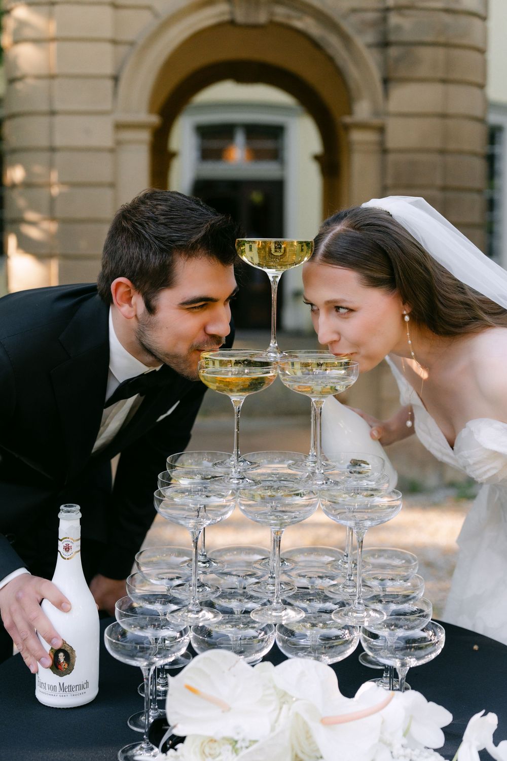 Wedding couple sips from a champagne tower display with stacked coupe glasses on a black table outdoors.