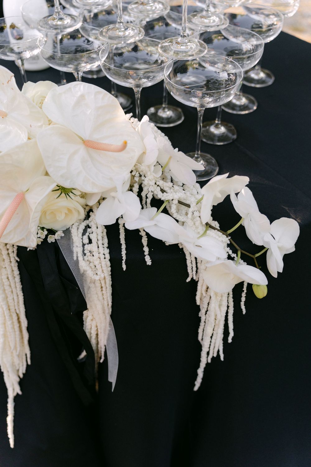 Elegant white floral arrangement with cascading orchids and crystal beads next to champagne glasses against black background.
