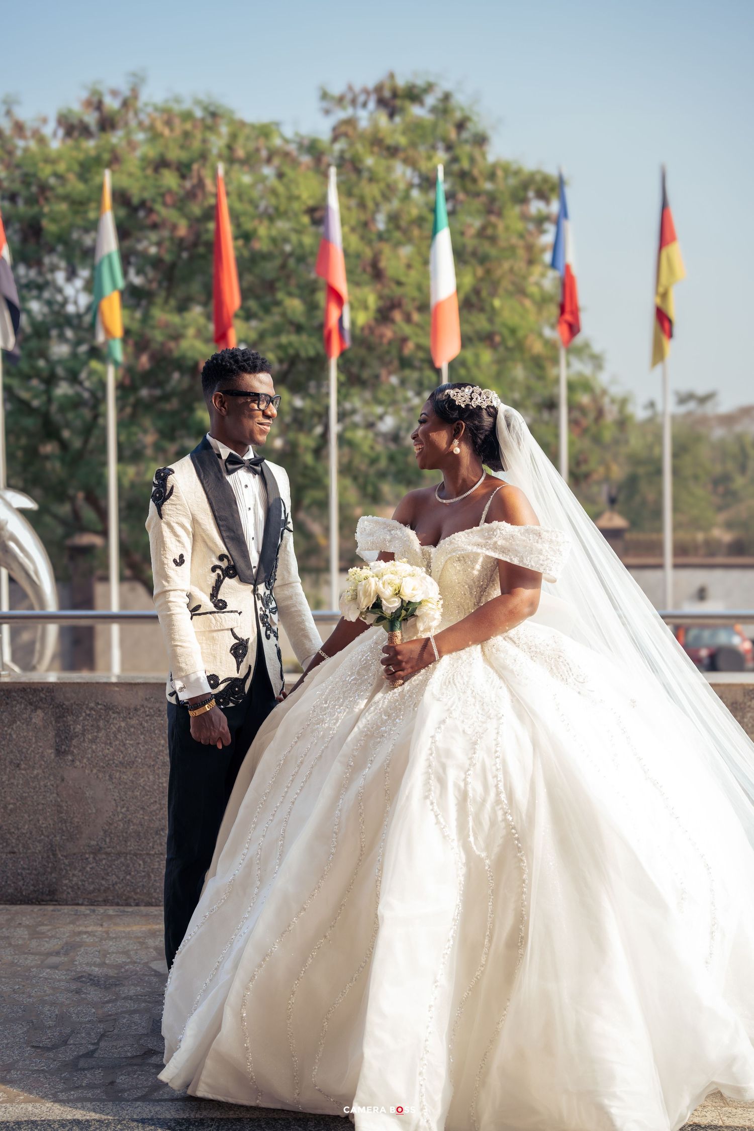 Wedding couple poses outdoors with international flags in background, bride in ball gown and groom in patterned tuxedo.