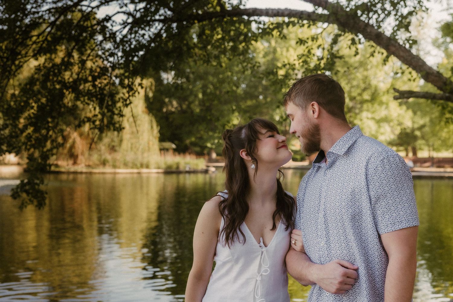 Two people in love share a romantic moment by a pond with trees in the background during golden hour. Photo & video engagement session.