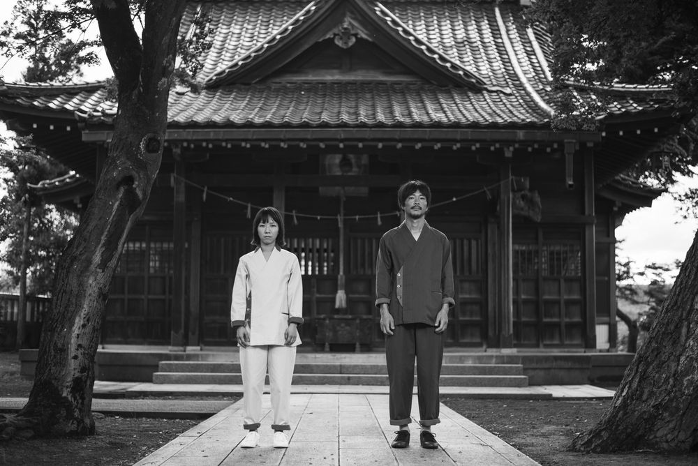 Two people stand in front of a traditional Japanese temple with curved roof and ornate architecture in black and white.